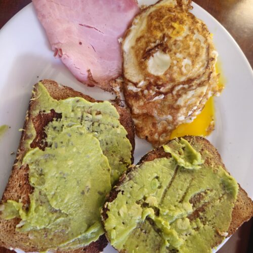 Two slices of whole wheat avocado toast served with crispy fried eggs and lean ham for a high-protein breakfast.